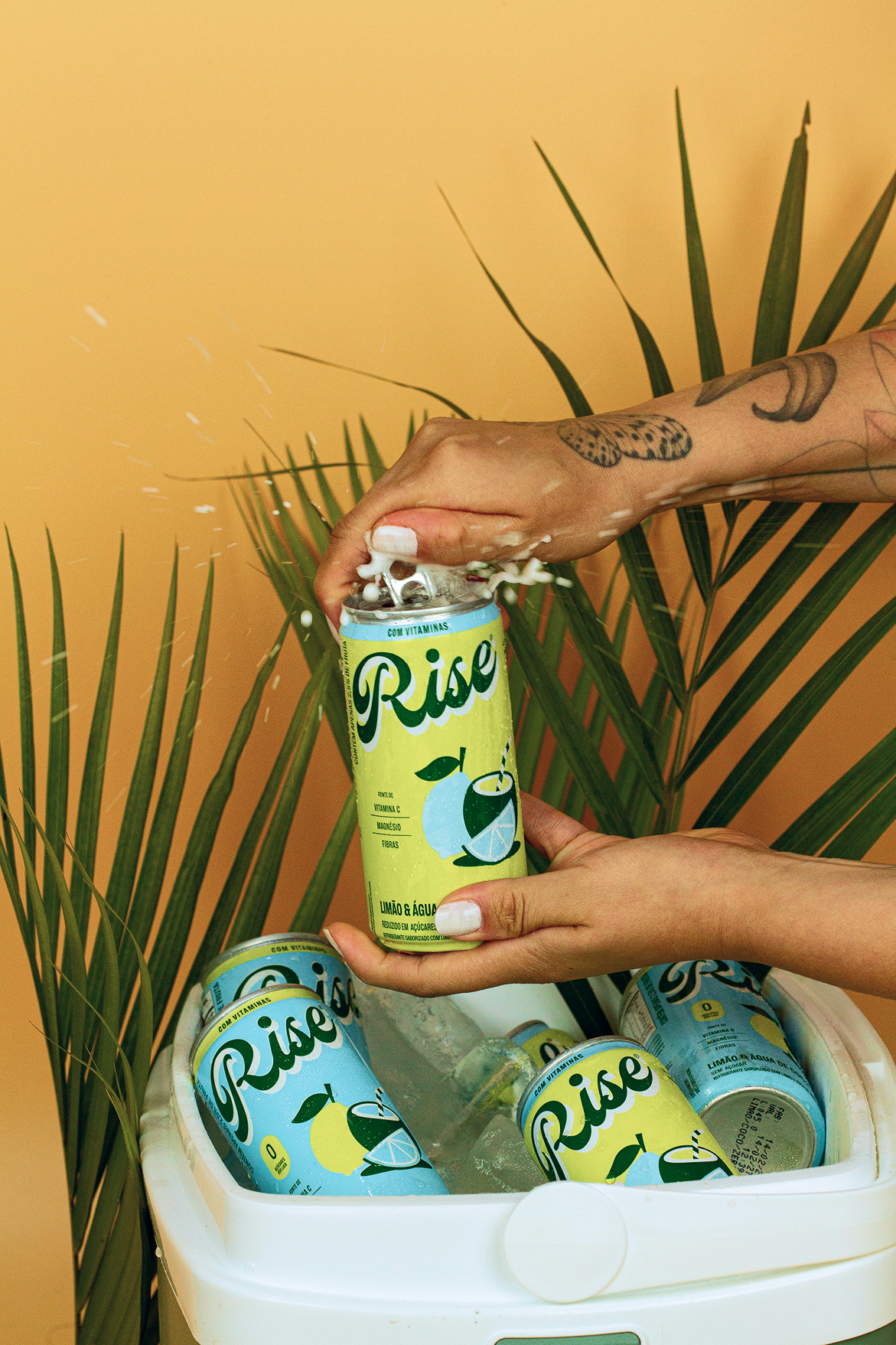 Mãos abrindo lata Rise Limão & Água de Coco com espuma, sobre cooler com gelo